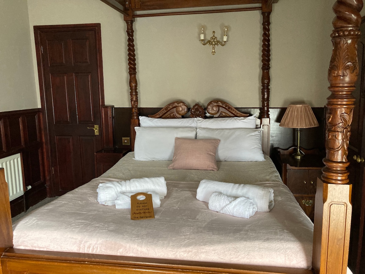 A carved four-poster bed occupies the center of the room, adorned with crisp white linens and a decorative pillow. Two rolled towels are neatly arranged at the foot of the bed. Soft natural light illuminates the space, highlighting the wooden furnishings and antique details.