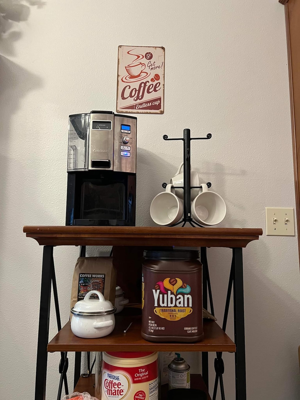 A coffee station is presented with a modern coffee maker featuring visible controls. Below, a wooden shelf holds a jar of Yuban coffee, a decorative white teapot, and two hanging coffee mugs on a wrought iron stand. Wall art depicting coffee is also displayed above.