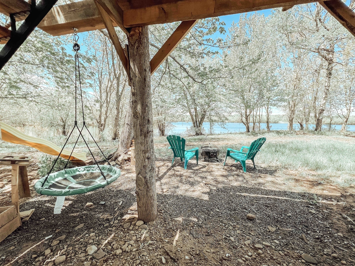 An inviting outdoor space features two green Adirondack chairs positioned around a firepit, with a swing chair hanging from a wooden structure. Soft grass and a scenic view of the water and trees create a peaceful setting.