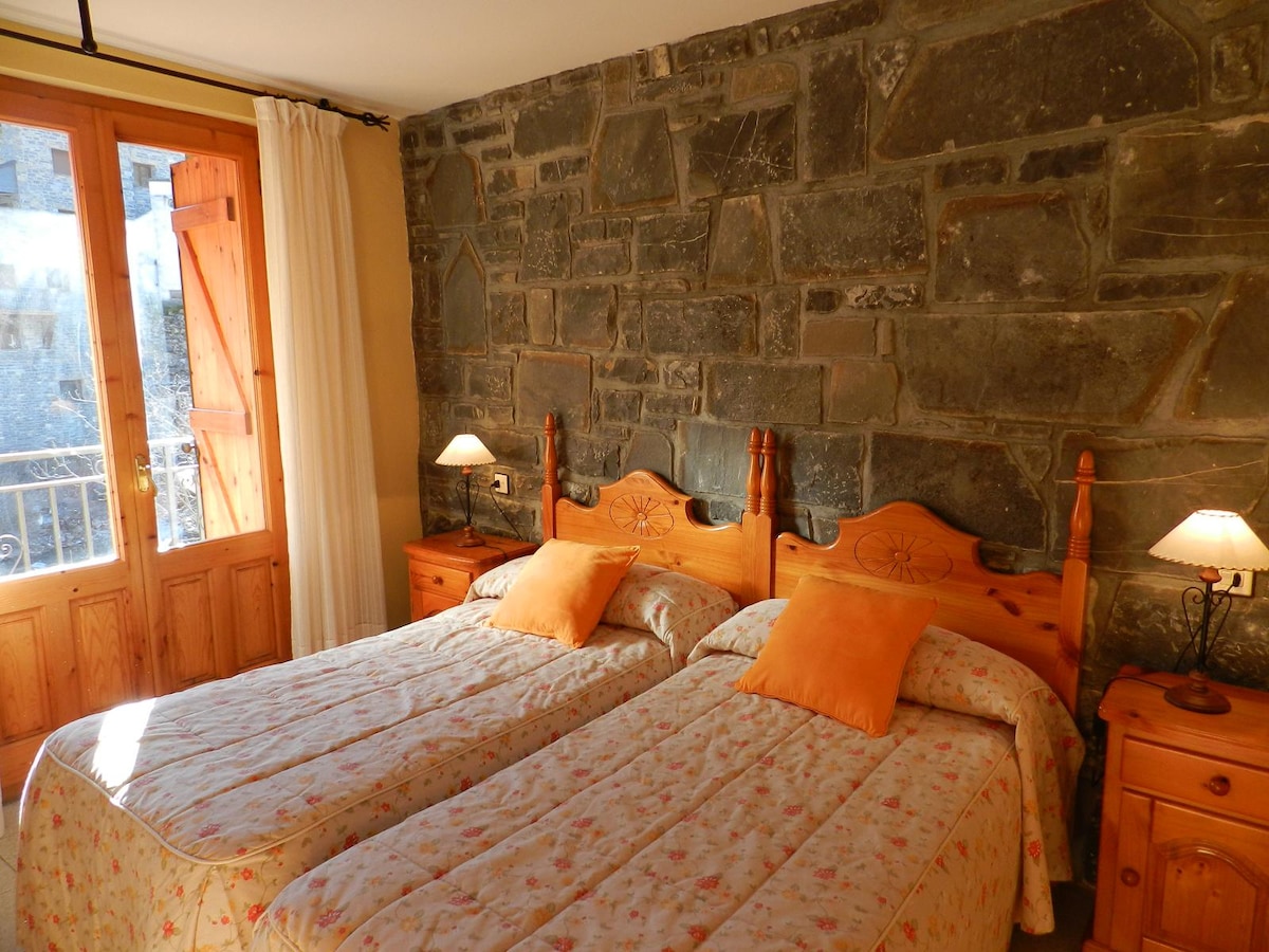 A room features two joined beds with floral bedding and orange cushions. The wooden walls are complemented by a stone accent wall. Bedside tables with lamps are positioned on each side. Natural light enters through a double door, leading to a small balcony.
