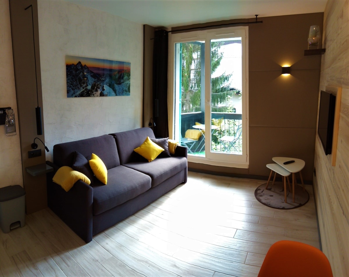 A living area is highlighted by a dark sofa adorned with yellow cushions. Large windows provide natural light and offer access to a small balcony. A round rug complements the space, and a small table is positioned beside the sofa, enhancing comfort and functionality.
