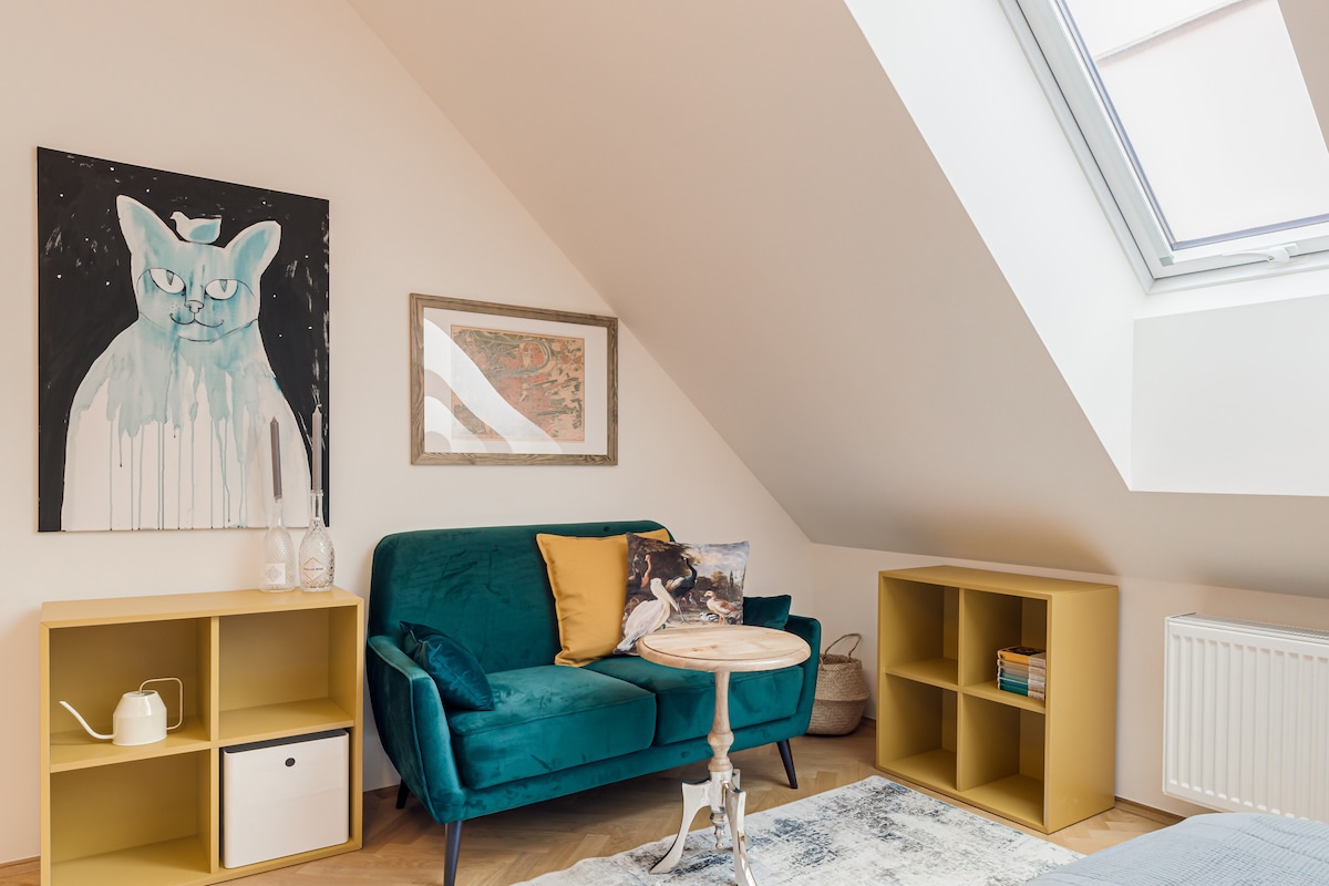 A bright corner of the apartment features a teal sofa adorned with decorative cushions. A small wooden table is positioned in front, accompanied by two yellow storage units. A framed artwork depicting a cat hangs above the sofa, while natural light streams in from a skylight.