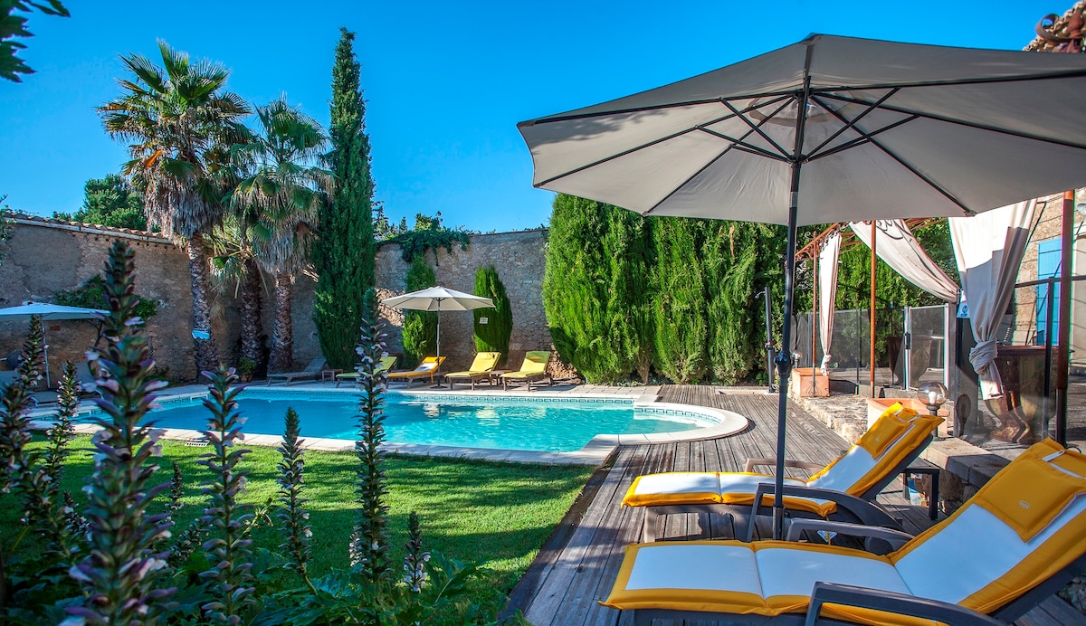 A shared swimming pool is surrounded by lush green gardens and tall trees, offering a serene environment. Sun loungers are positioned beside the pool, shaded by large umbrellas. The area features wooden decking and a mix of vibrant flowers, creating a refreshing outdoor space.