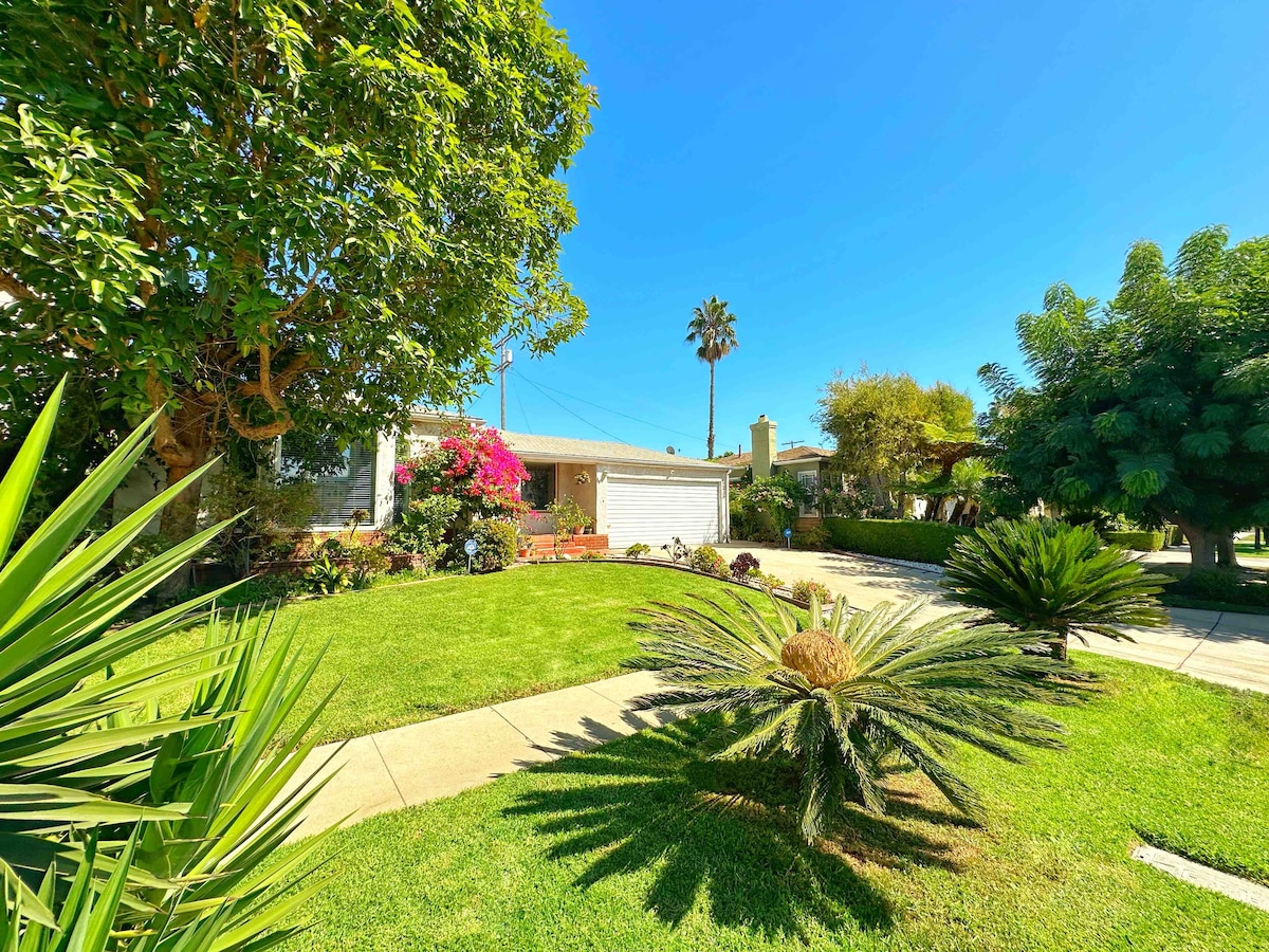 A well-kept front yard is surrounded by lush greenery, featuring palm and succulent plants. The vibrant blooms of bougainvillea add color against a clear blue sky. A driveway leads to a garage, and a relaxing atmosphere is highlighted by the neatly trimmed lawn.