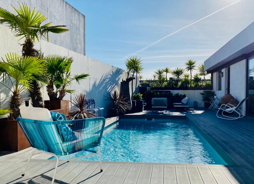 A sunlit pool area is surrounded by tropical plants, creating a serene atmosphere. The swimming pool is framed by gray decking, with lounge chairs positioned nearby for relaxation. Bright blue accents on seating provide a refreshing touch against the neutral backdrop.