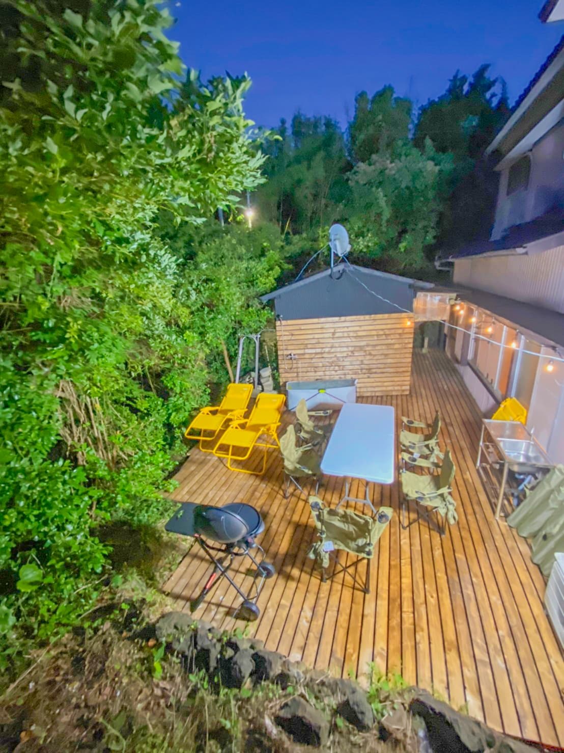 A wooden deck is adorned with yellow chairs and a large table, surrounded by greenery. A barbecue grill is positioned nearby, and string lights add a soft glow above. The deck provides an inviting space for outdoor gatherings, set against a nighttime backdrop.
