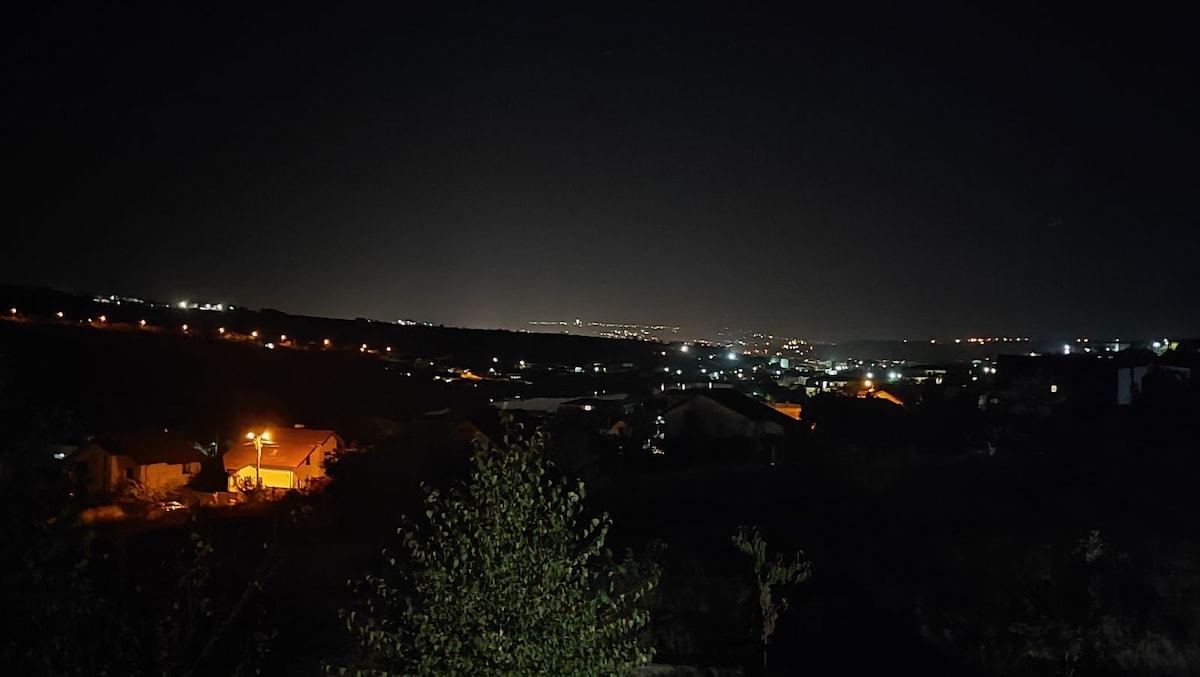 A nighttime view showcases a valley illuminated by a spread of soft lights from homes and streets. The distant horizon reveals a subtle glow, while the foreground features a silhouette of trees, adding depth to the scenic landscape.