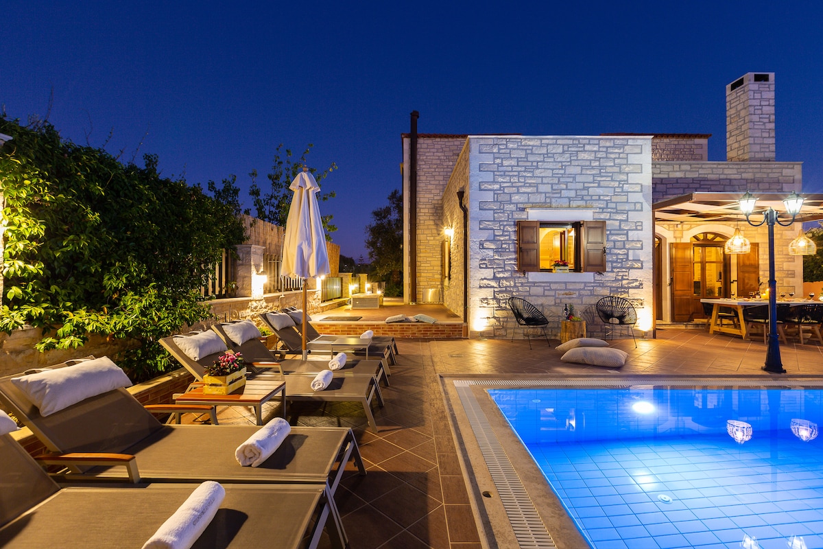 The outdoor area features a private swimming pool illuminated in blue, surrounded by eight sunbeds with rolled towels. A dining table and chairs are situated under a wooden pergola, with a few decorative plants enhancing the tranquil environment. Soft lighting creates a serene evening setting.