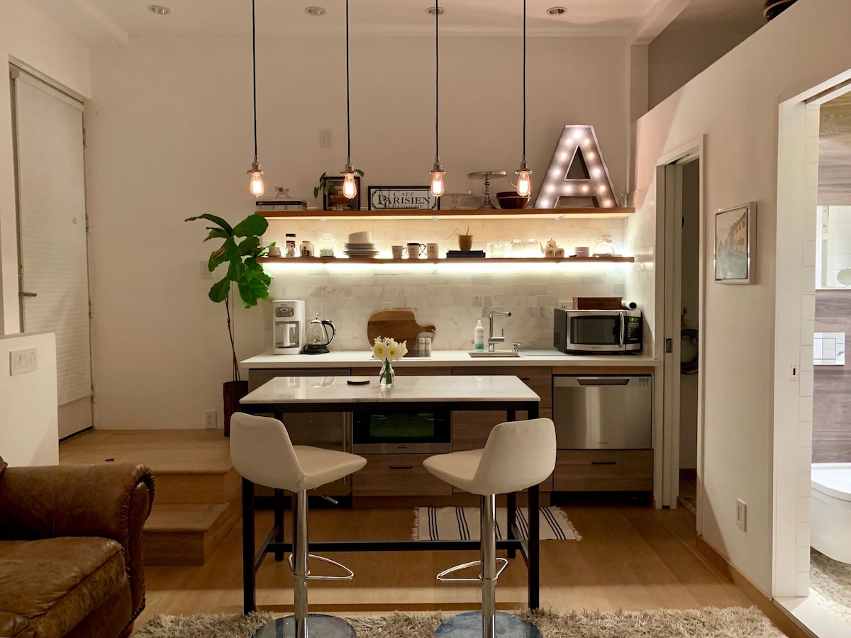 A modern kitchenette is displayed, featuring sleek cabinets and high-end appliances such as a microwave and coffee maker. A small dining area is complemented by two bar stools. Soft lighting highlights the decor, including a decorative plant and a vase with flowers on the countertop.