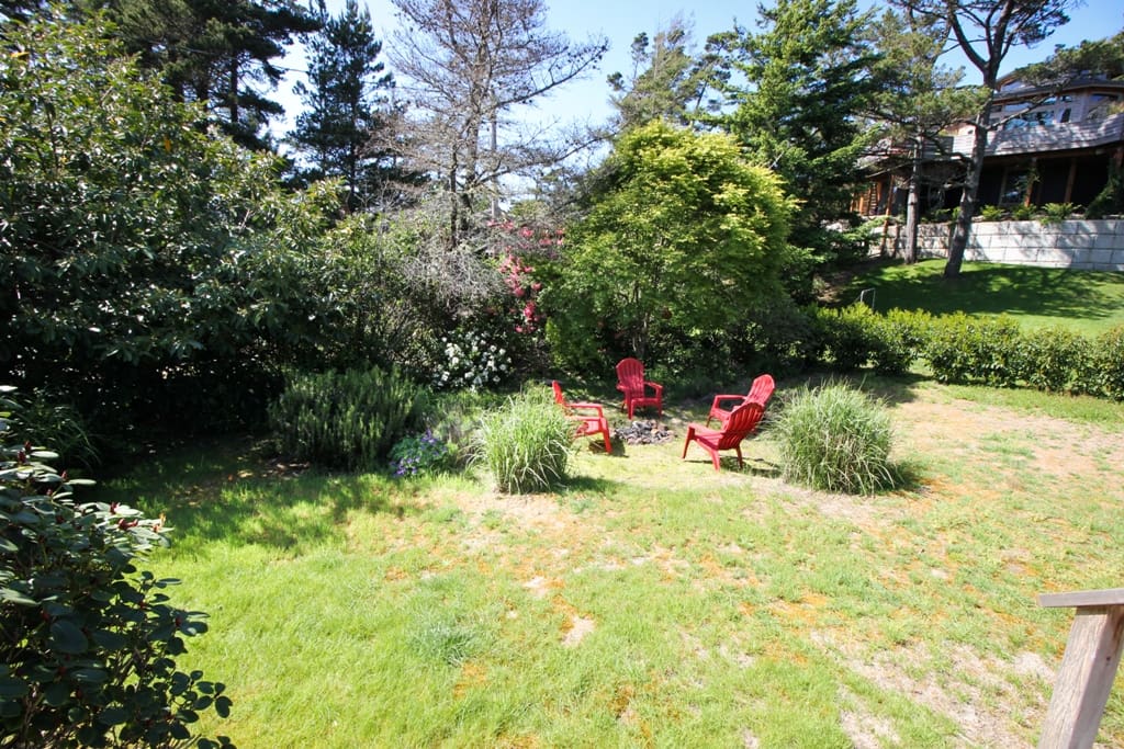 Large grassy landscaped backyard with fire pit and deck with table, chairs and charcoal BBQ.