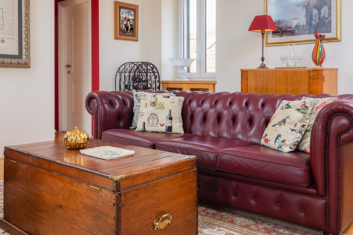 A deep red leather sofa with decorative pillows is positioned in a well-lit living area. A rustic wooden coffee table sits in front, complemented by a patterned area rug. Natural light streams in through large windows, enhancing the comfortable and inviting space.