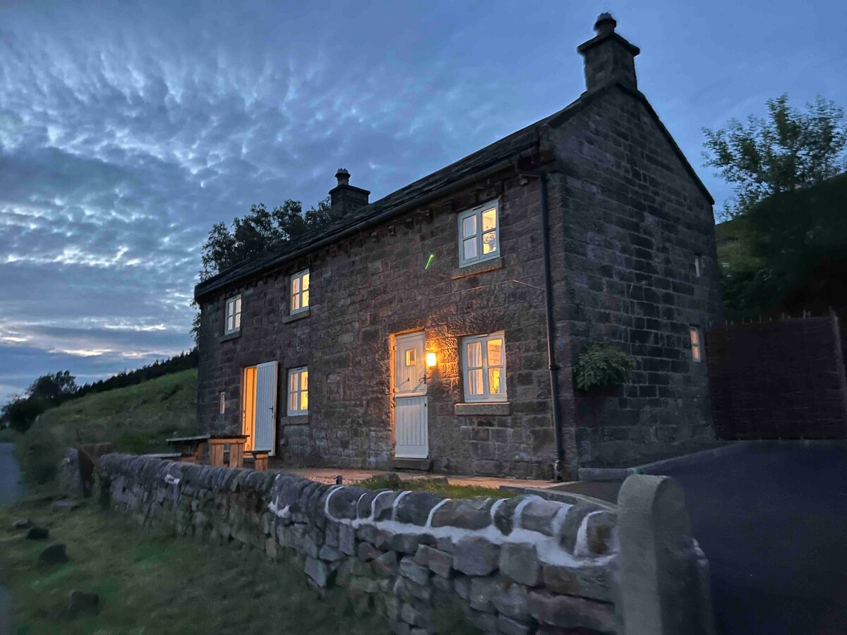 The stone exterior of Roachside Cottage is softly illuminated by warm light, highlighting the charming windows and door. The surrounding landscape features rolling hills under a subtly clouded sky, creating a tranquil evening atmosphere. A stone wall borders the property, adding character to the setting.