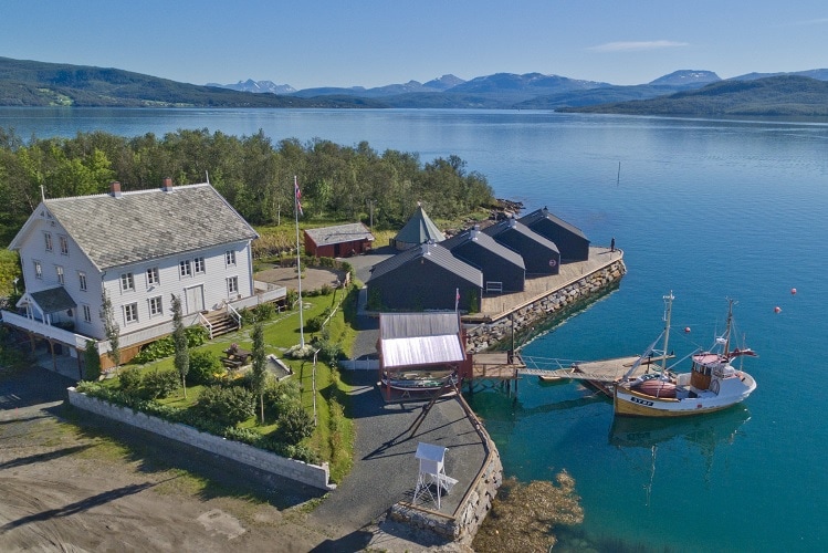 An aerial view captures the charming Finsnes Gaard alongside a serene waterfront, featuring traditional wooden buildings and a bright fishing boat moored by the dock. The surrounding landscape showcases calm waters and distant mountains, creating a tranquil setting.