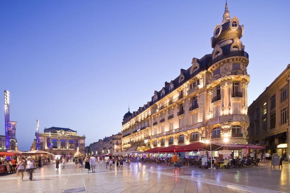 A vibrant urban square is captured during twilight, showcasing illuminated historic buildings and lively cafes with outdoor seating. People can be seen strolling leisurely across the open space, while decorative lights enhance the evening ambiance.