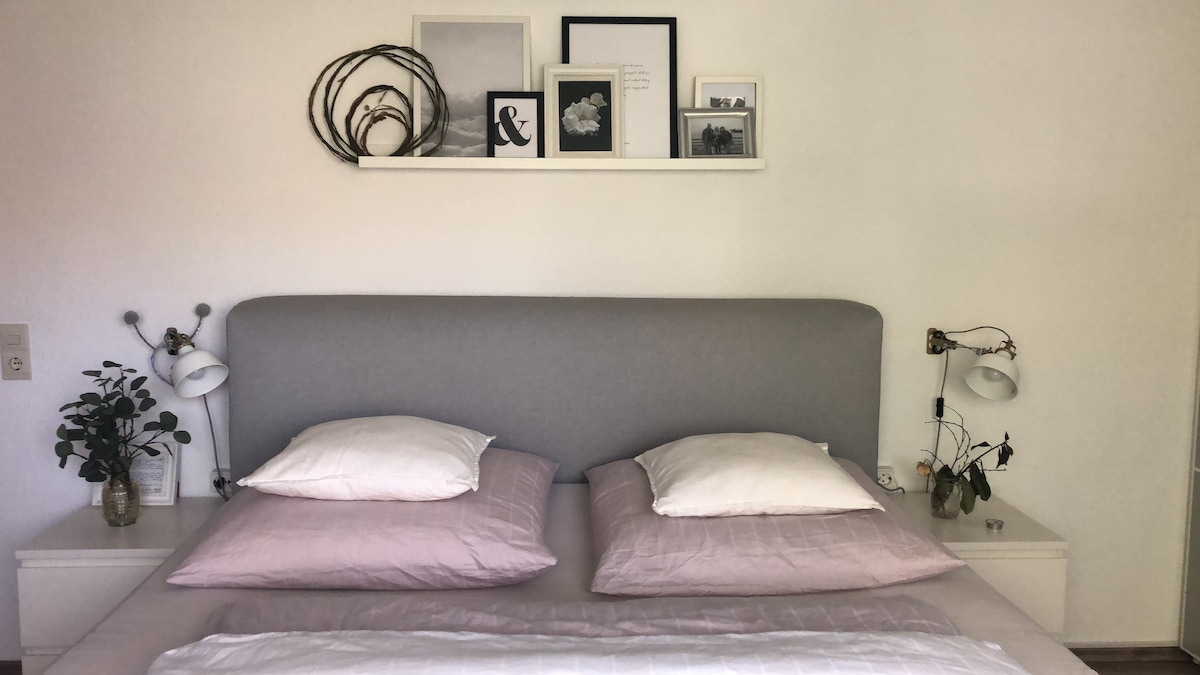 A spacious bed is centered against a light-colored wall, featuring soft gray and neutral bedding. Framed images and decorative objects are displayed on a shelf above the headboard. Bedside lamps and a small plant add subtle touches to the serene bedroom setting.