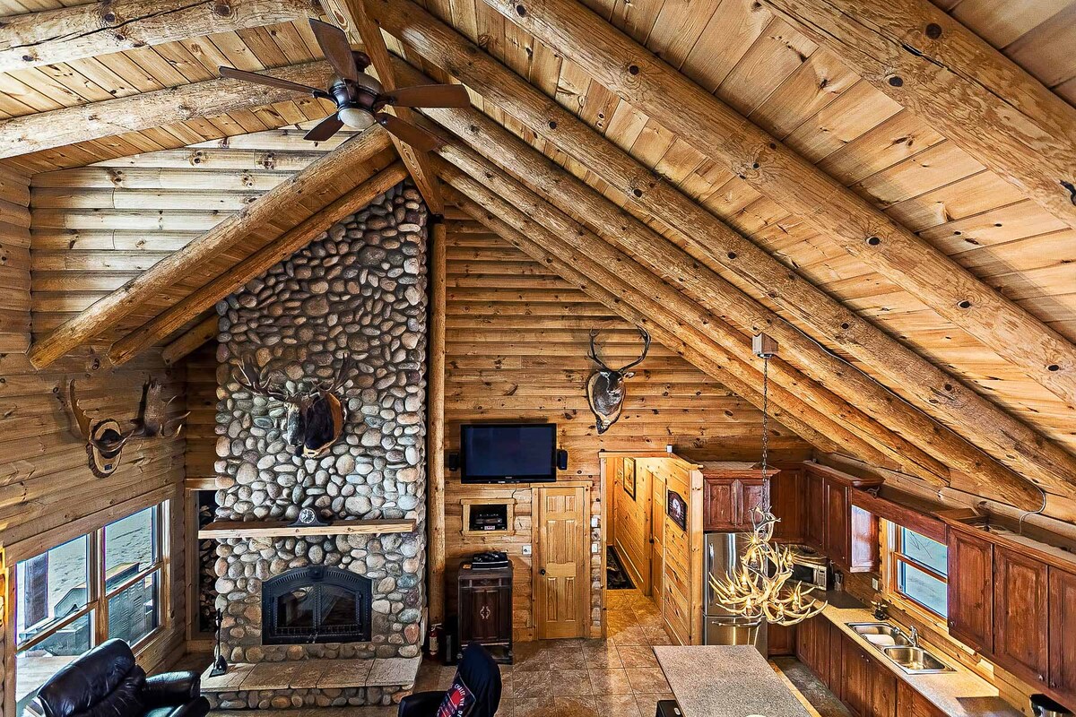 The spacious living area features exposed wooden beams and stone accents, highlighted by a large fireplace. Rustic decor, including mounted trophies, adorns the walls. A central island with seating allows for social gatherings, while large windows offer natural light and views of the surrounding nature.