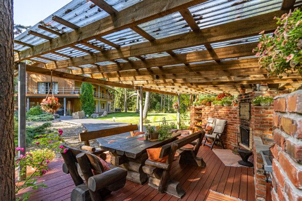 An outdoor dining area is shown, featuring a large wooden table surrounded by several benches. A pergola offers partial shade, and potted flowers add touches of color to the environment. The green landscape can be seen in the background, enhancing the natural setting.