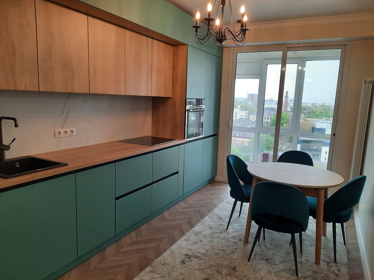 A modern kitchen features light wood cabinetry with a green accent. A round dining table with four blue chairs is positioned on a textured rug, while large windows provide natural light and a view of the city skyline.