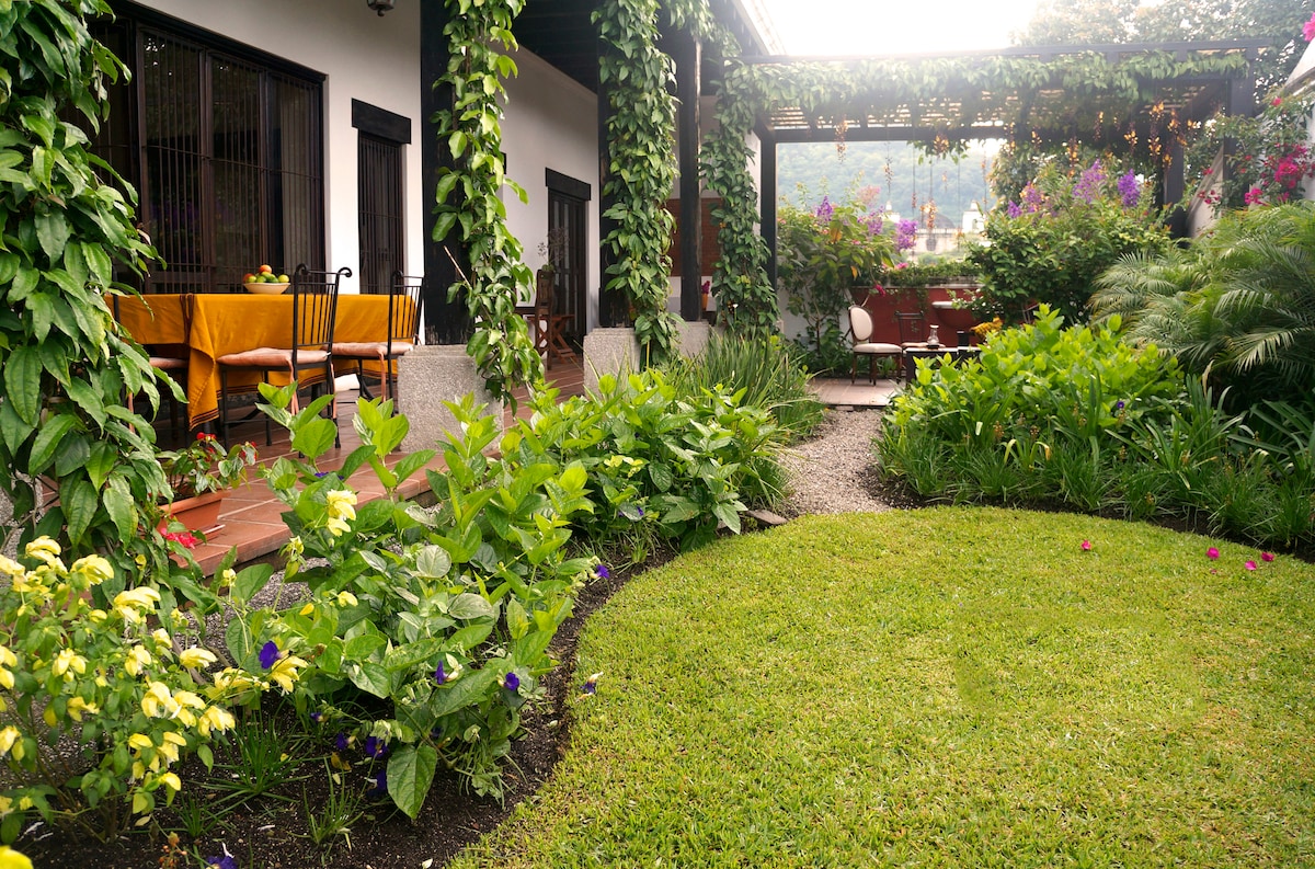 A lush garden surrounds a patio area, featuring vibrant green grass and colorful flowers. Climbing vines adorn the walls of the entrance, while a dining table with chairs is set under a shaded pergola, creating an inviting outdoor space.