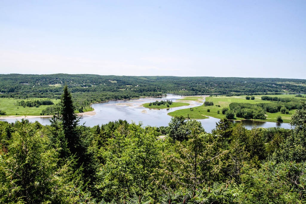 A panoramic view of lush green hills and winding rivers is captured from an elevated vantage point. The landscape features a mix of foliage and open fields under a clear blue sky, highlighting the tranquil beauty of the natural surroundings.