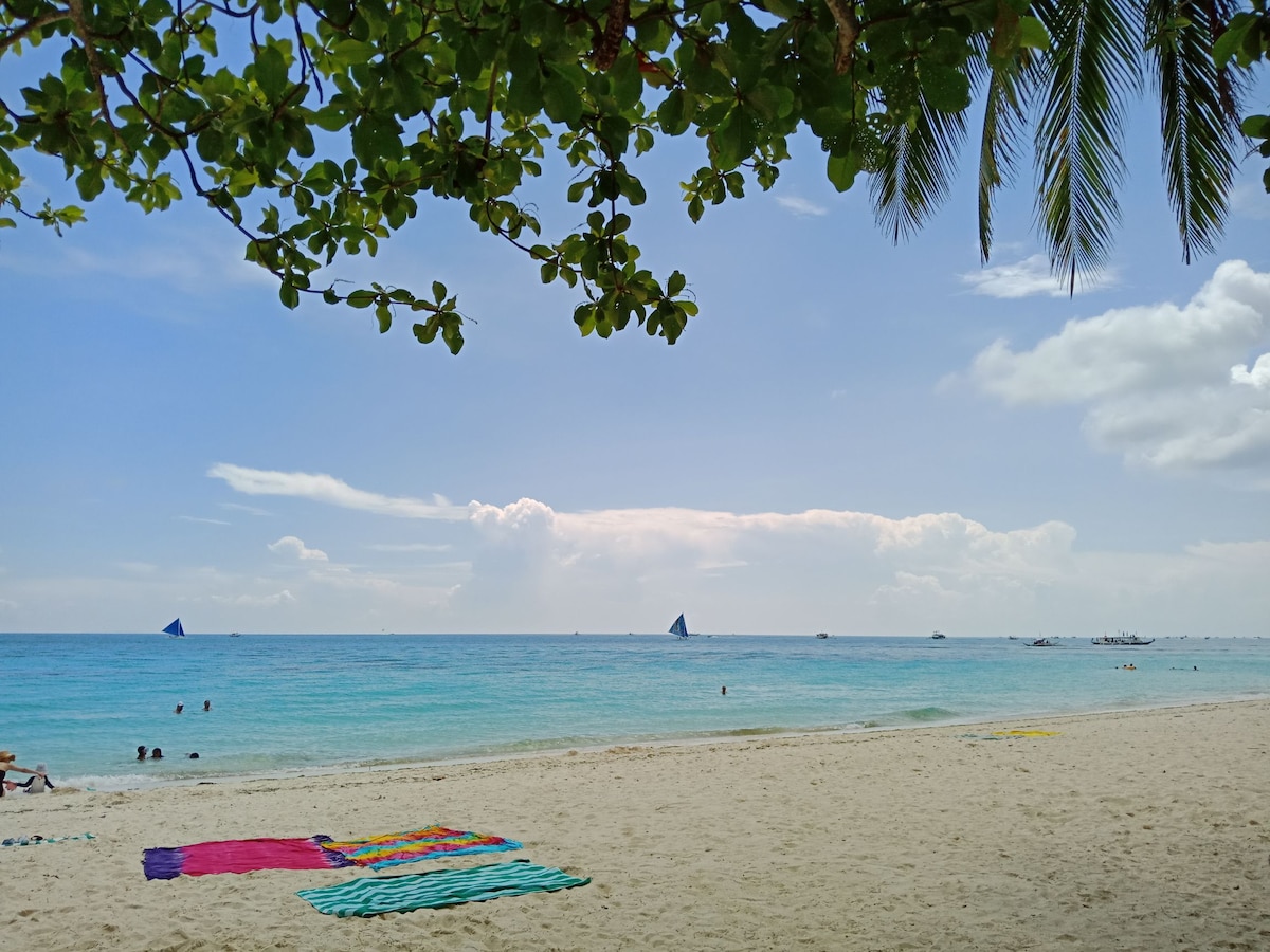 A sandy beach is depicted under a bright sky, with gentle waves lapping at the shore. Colorful beach towels are spread out on the sand, while small boats are visible on the horizon. Lush green leaves frame the top of the image.