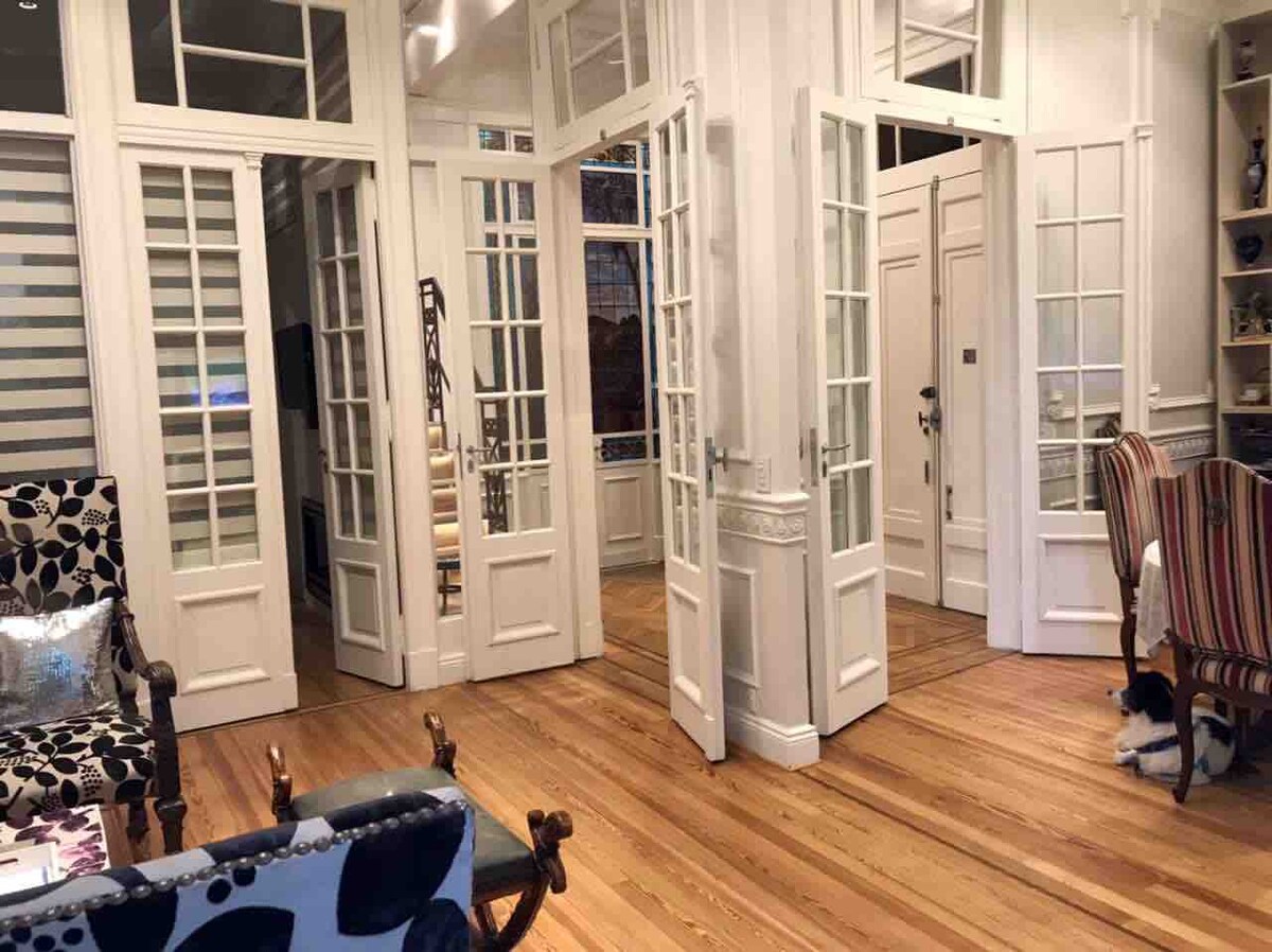 Multiple glass-paneled doors create a series of elegant entries, leading into a well-lit interior. Rich wooden floors complement the stylish design, while strategically placed furnishings enhance the room's spaciousness. Natural light filters in, accentuating the inviting atmosphere of the space.