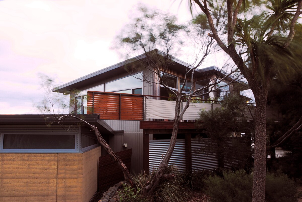 The exterior of the residence features a combination of natural materials, including rammed earth and timber. Large windows allow for ample natural light, while a deck with wooden slats offers outdoor space. Surrounding greenery contributes to a peaceful, natural setting.