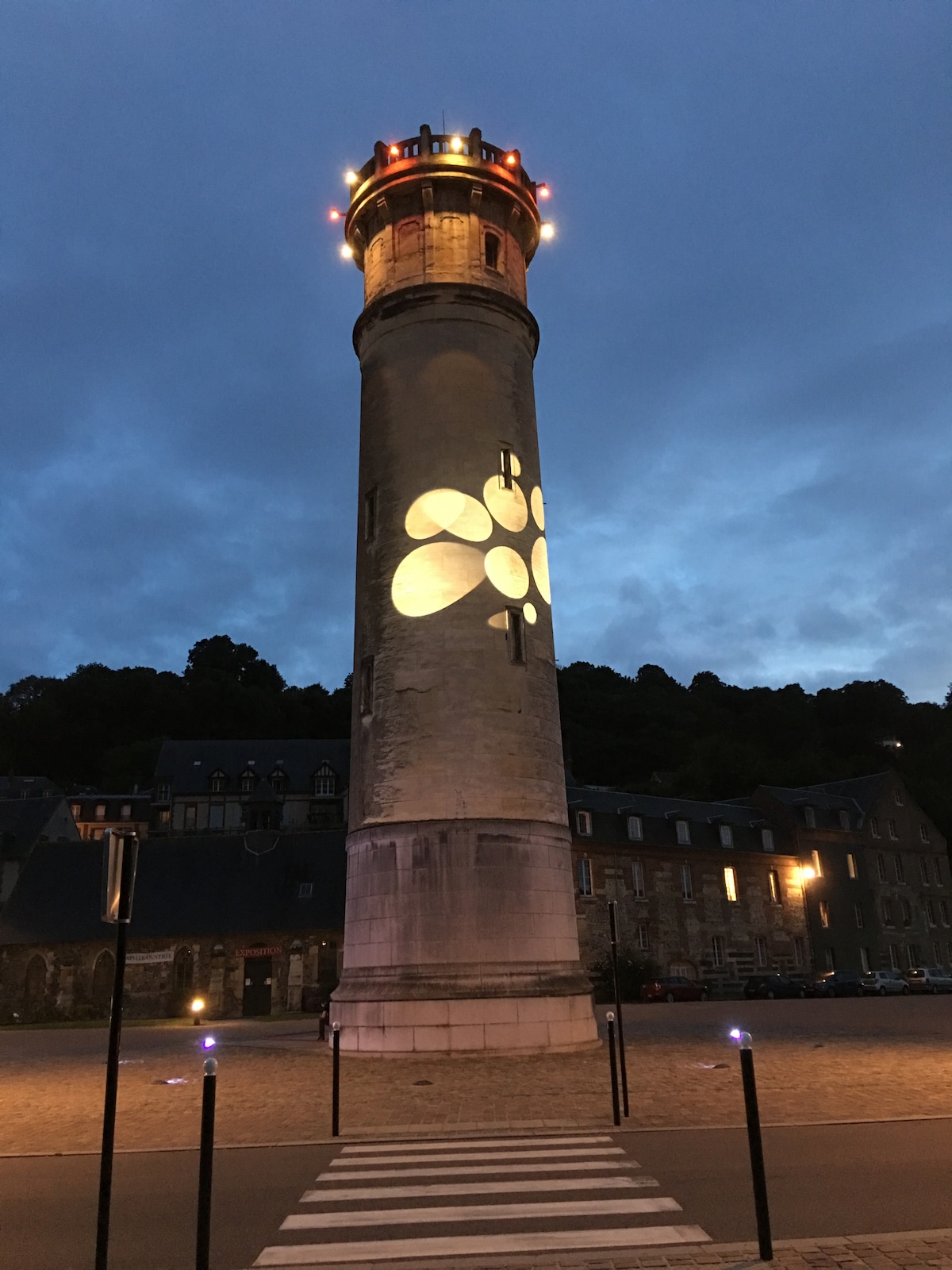 A historic lighthouse stands tall against a twilight sky, illuminated with soft lights projecting floral patterns onto its stone surface. The surrounding area features a cobblestone path and buildings, creating an atmosphere of tranquility in this quiet location.