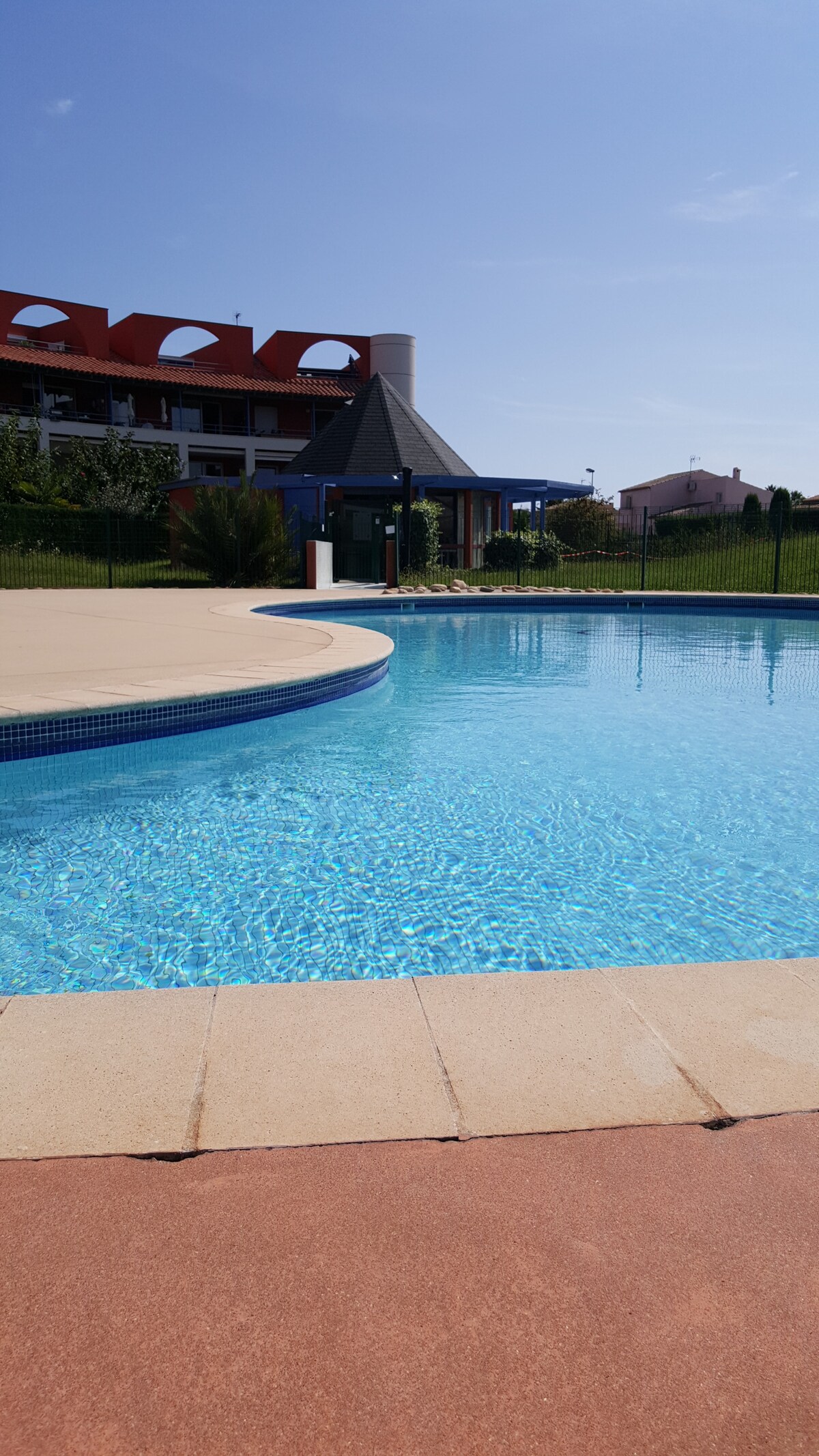 A clear blue swimming pool is surrounded by a curved tan-colored deck. Lush green landscaping is visible beyond the pool, with a red-roofed building in the background. The sky above is bright and sunny, contributing to a welcoming atmosphere.