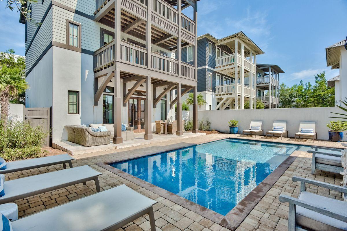 A spacious outdoor area surrounds a private heated pool, with comfortable lounge chairs arranged poolside. The three-story home is visible in the background, framed by greenery and offering multiple outdoor living spaces. Clear blue skies reflect on the water, enhancing the tranquil setting.