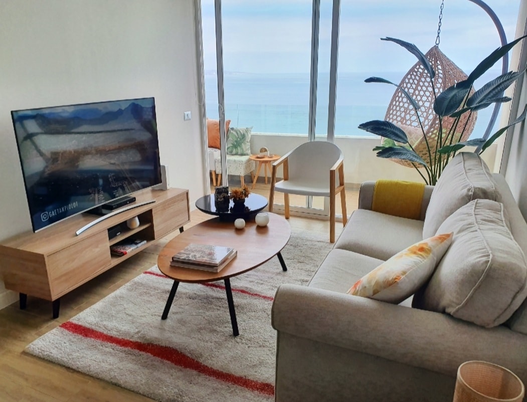 A light-filled living area features a comfortable sofa and a stylish coffee table with decorative accents. A large TV is positioned on a modern media console, while a hanging chair adds a touch of relaxation. Panoramic views of the sea are visible through expansive windows.