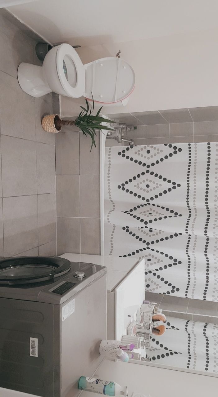 A modern bathroom is presented with neutral grey tiles. A white toilet is visible alongside a bathtub with a patterned shower curtain. A laundry machine is positioned in the space, and a potted plant adds a touch of greenery. Various toiletries are organized on the countertop.