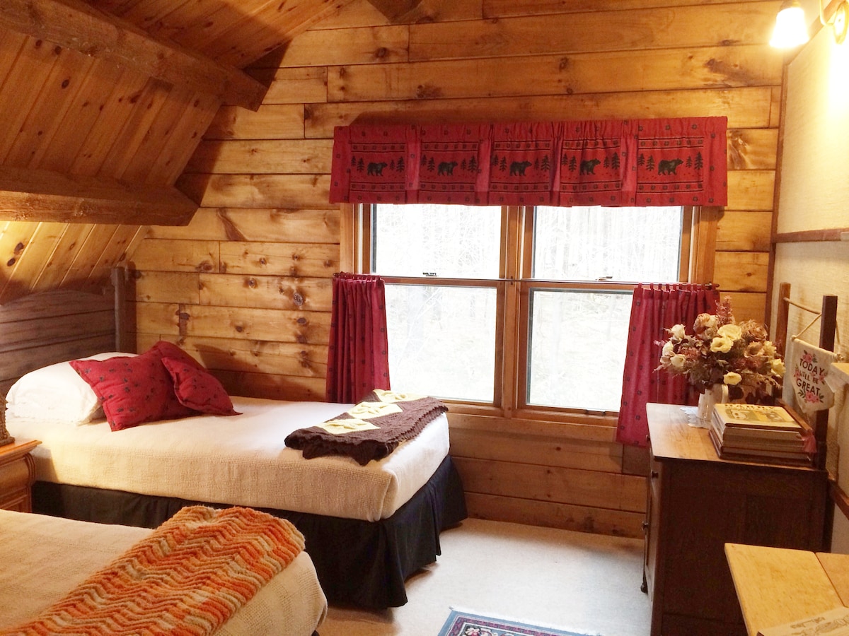 A cozy rustic bedroom features two twin beds, each with white linens and dark bed skirts. Natural wood walls provide warmth, complemented by red valances over the windows. A dresser and a small bookshelf are positioned beside the beds, adding functionality to the inviting space.