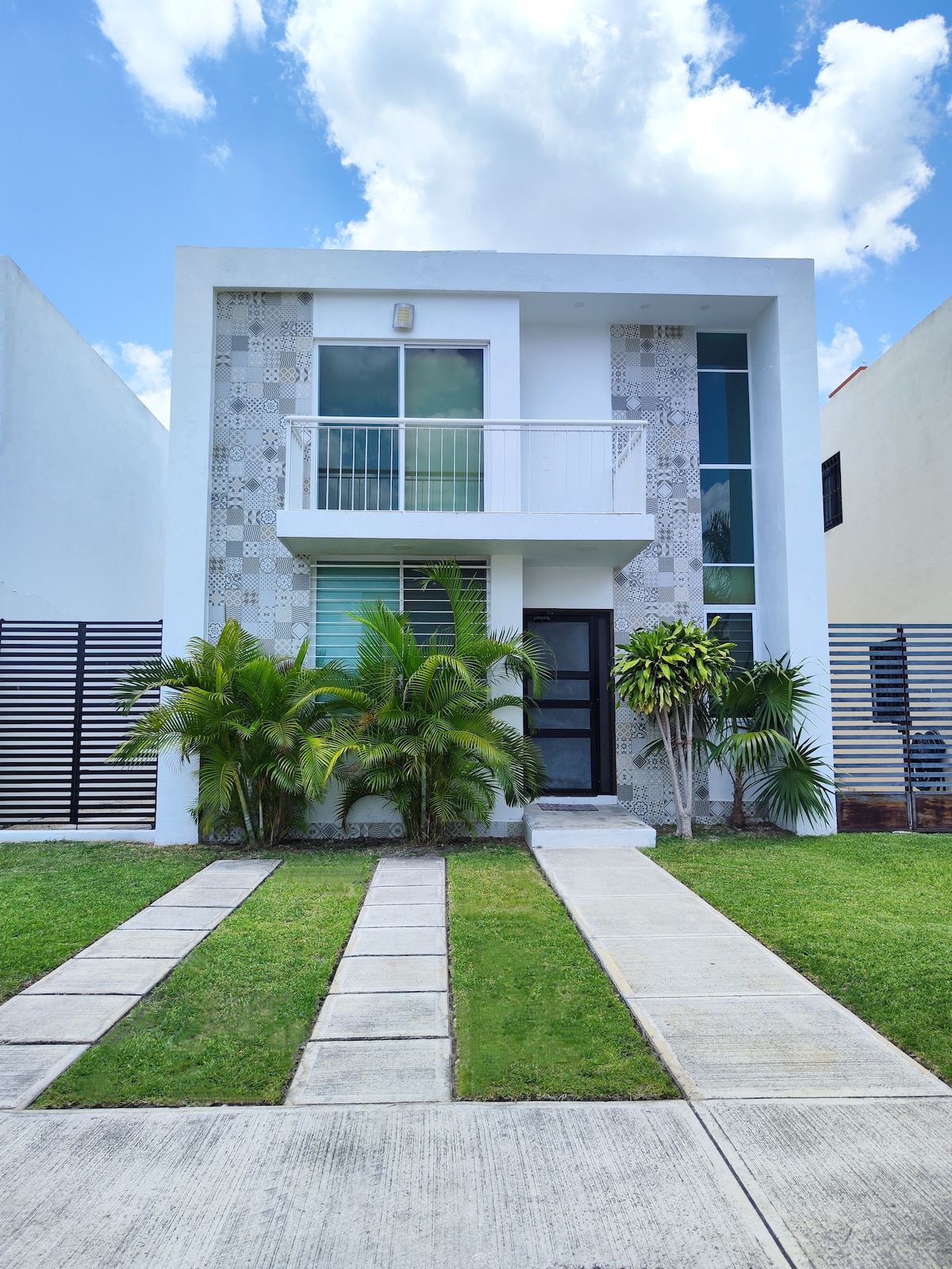 The two-story house features a modern facade with decorative elements and large glass windows. A walkway lined with tropical plants leads to the front door. The well-maintained lawn is complemented by neatly paved pathways, reflecting a clean and inviting exterior.