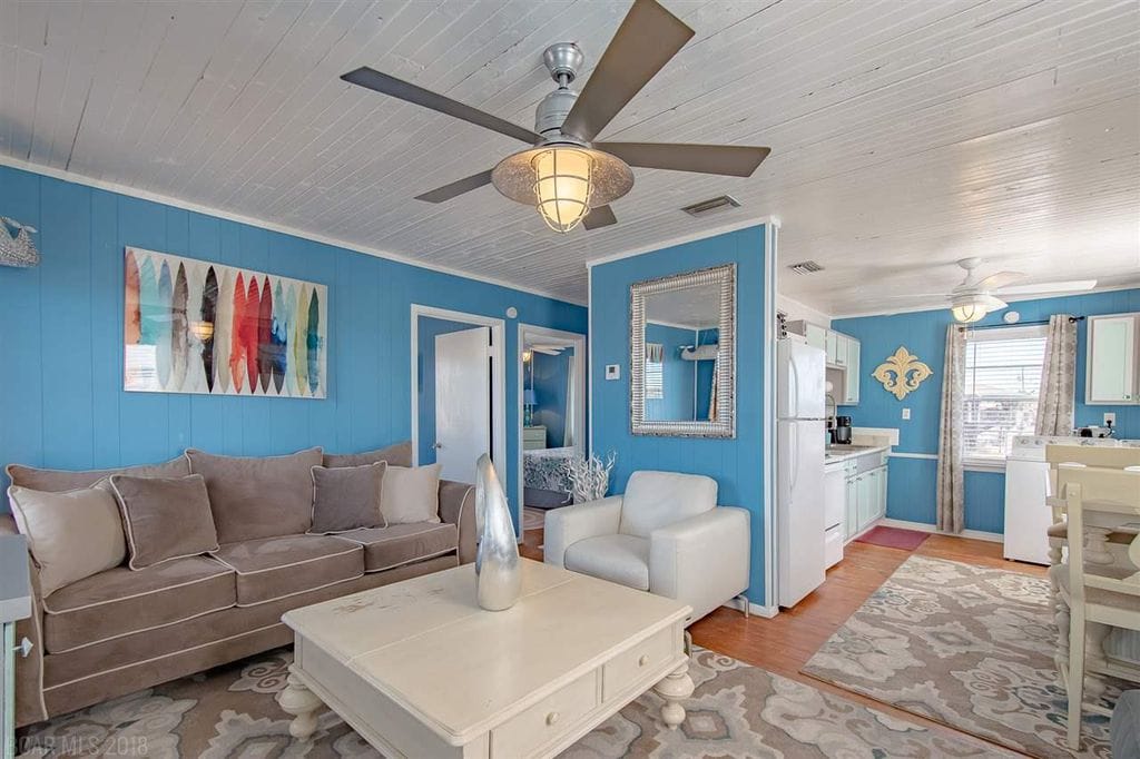 A bright living area features a comfortable sectional sofa and a white armchair, complemented by a decorative coffee table. A ceiling fan hangs above, and the walls are painted in a calming blue hue. A dining space is visible in the background.