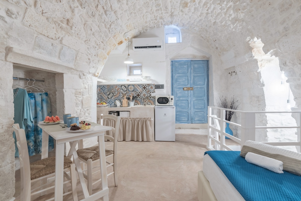 A charming interior features a light-colored stone archway, enhancing the rustic appeal. A small dining table with two chairs is set beside a kitchenette, while a comfortable sleeping area is adorned with a blue blanket. Bright blue wooden doors add a pop of color.