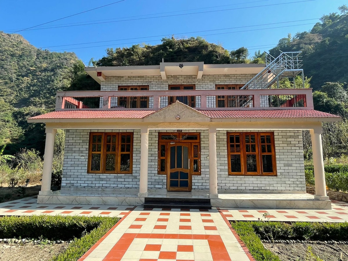 The house features a stone exterior with wooden window frames and a red tiled roof. A spacious front porch is flanked by neatly arranged flower beds, while a pathway leads to the entrance. Surrounding hills add a serene backdrop.