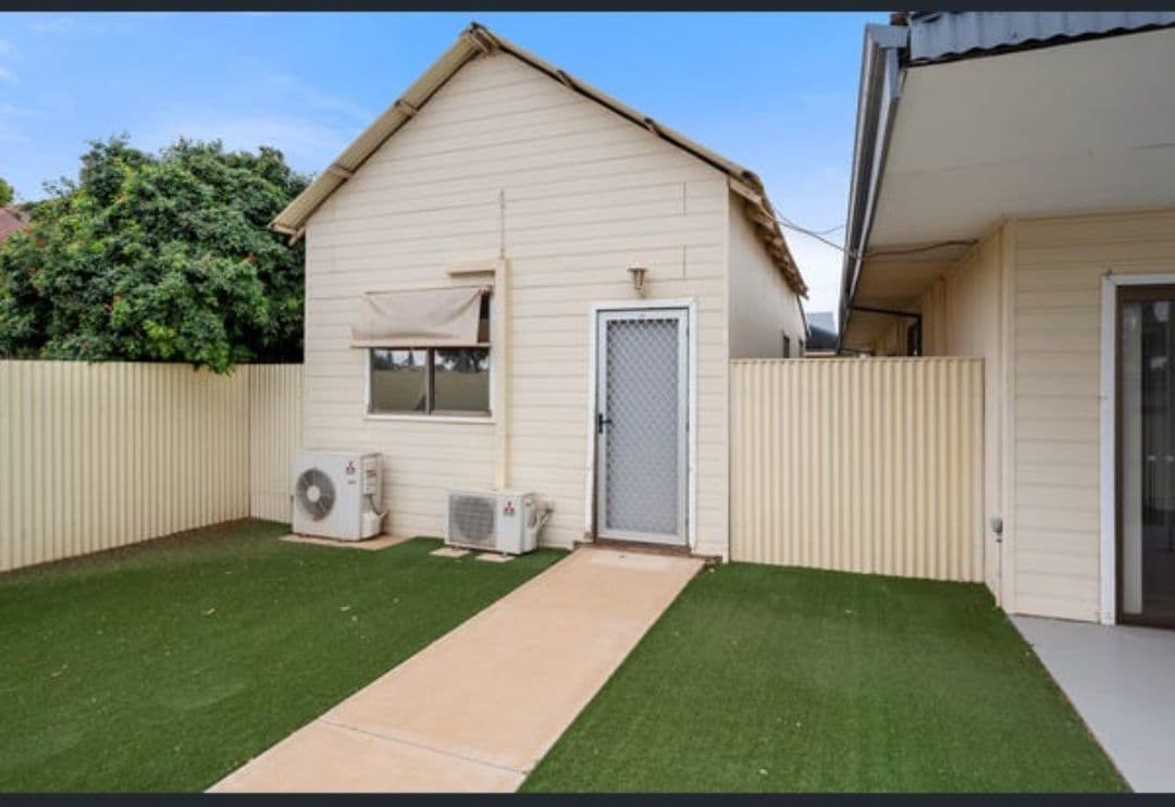 An exterior view of a charming granny flat features a well-maintained lawn, bordered by beige fencing. An entrance door with a security screen is visible. Two air conditioning units provide comfort, while a clear pathway leads to the entrance, surrounded by greenery.