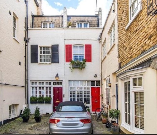 The exterior of the mews house is displayed, featuring a charming façade with white walls and contrasting dark shutters. Bright red doors are framed by potted plants. A silver car is parked in front, surrounded by cobblestone and welcoming residential architecture.
