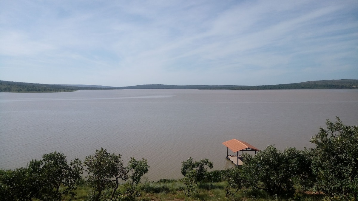 A wide view of a calm lake is captured, featuring soft ripples under a blue sky. The horizon is lined with greenery, and a small dock with a roof stands at the water's edge, providing access to the serene landscape.