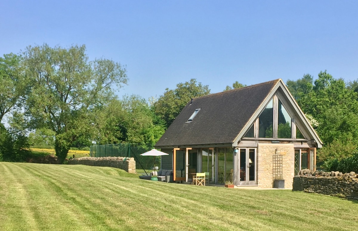 The bespoke oak building is positioned on a spacious lawn, framed by greenery and trees. Large windows allow for abundant natural light, while a private patio area features a parasol and bistro table. A nearby tennis court may also be visible.