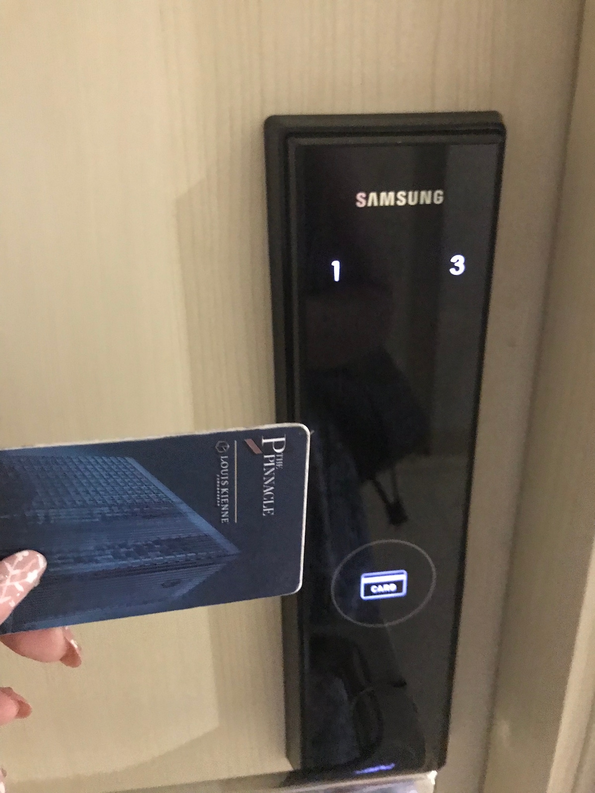 A modern electronic door lock is displayed, featuring a sleek black panel with illuminated numbers. A hand is shown holding a key card in front of the lock, ready for use. The overall design emphasizes security and convenience.