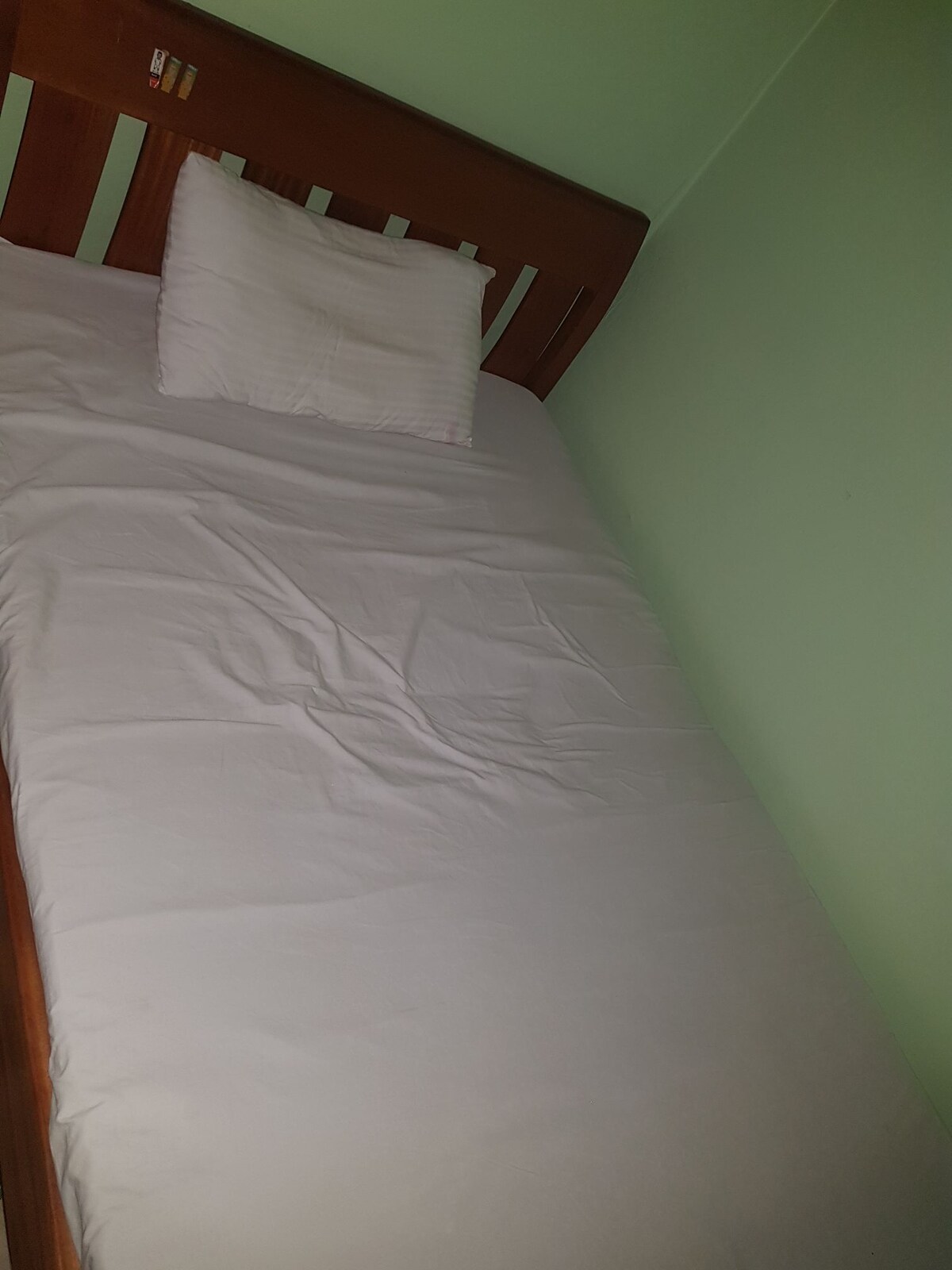 A single bed is positioned against a light green wall, featuring a simple wooden headboard and a soft, white blanket. A small, white pillow is placed at the head of the bed, providing a neat and comfortable appearance.