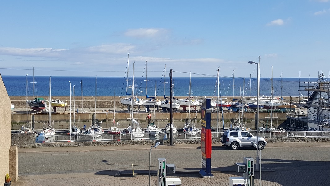 A view of the west harbour marina is presented, featuring several moored sailboats and yachts. The peaceful sea stretches into the distance under a clear blue sky, with light clouds visible. A vehicle is parked nearby, adding to the coastal scene.