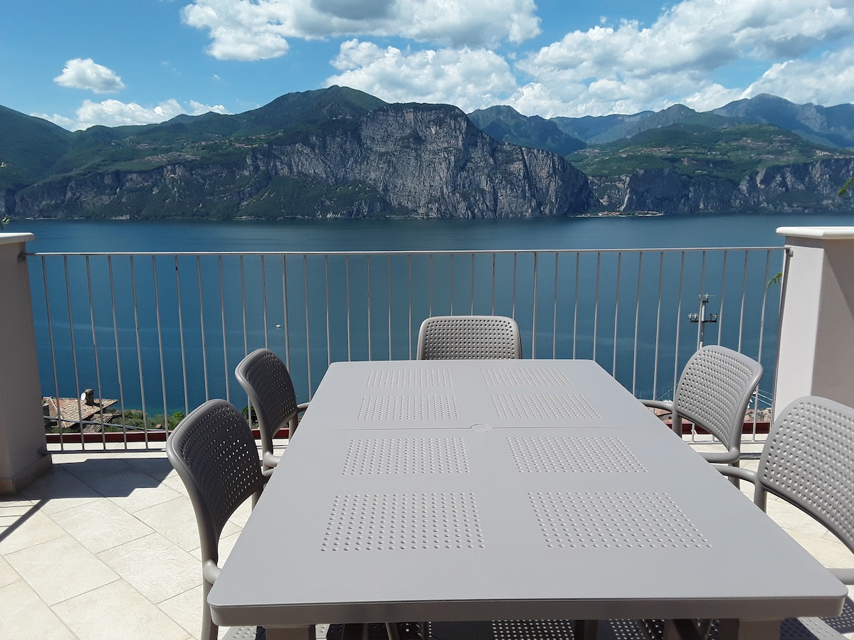 A spacious terrace features a large rectangular table surrounded by six chairs. The view beyond includes a serene lake and dramatic mountain scenery under a partly cloudy sky.