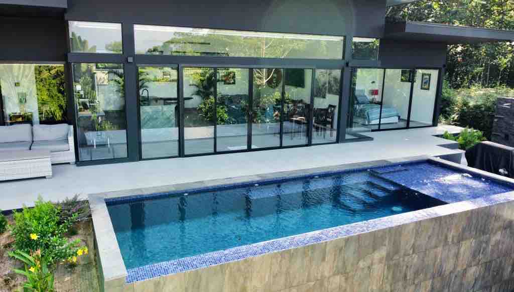 An infinity pool is situated in front of a modern house, featuring large glass doors that provide views of the surrounding jungle. Tropical greenery is visible, enhancing the serene environment. A seating area with sofas is positioned nearby, offering a place to relax.