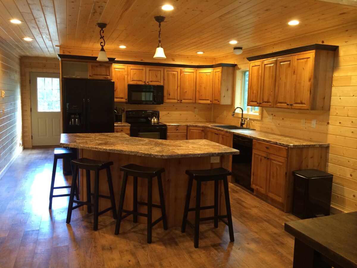 The kitchen features wooden cabinetry and a spacious island with bar stools. Modern appliances are included, with a black refrigerator and stove contrasting against the warm wood tones. Natural light streams in through a nearby door, enhancing the inviting atmosphere.