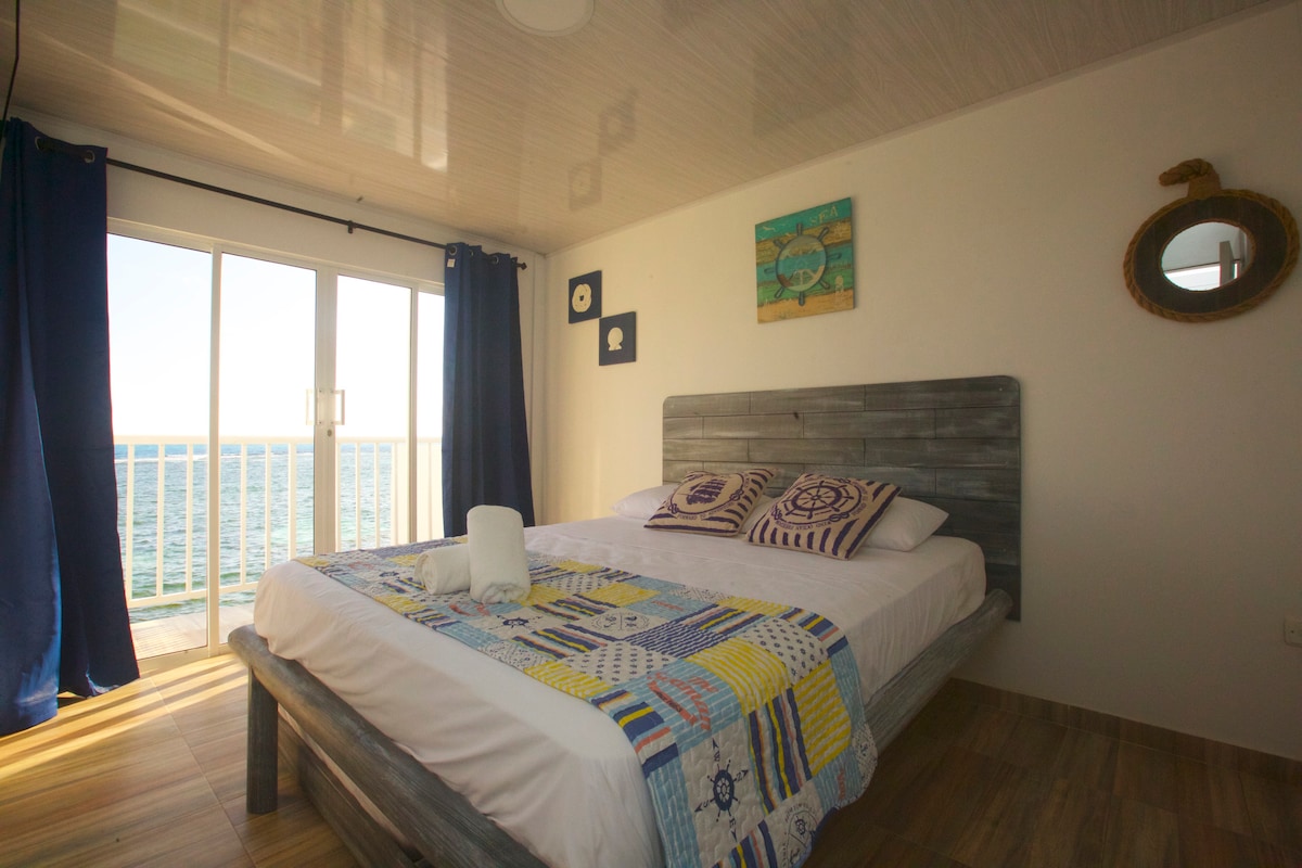 A bright sleeping area features a spacious bed adorned with a colorful quilt. Soft natural light filters through large glass doors, revealing a view of the ocean. Dark blue curtains provide contrast against the light walls, complemented by nautical-themed decor.