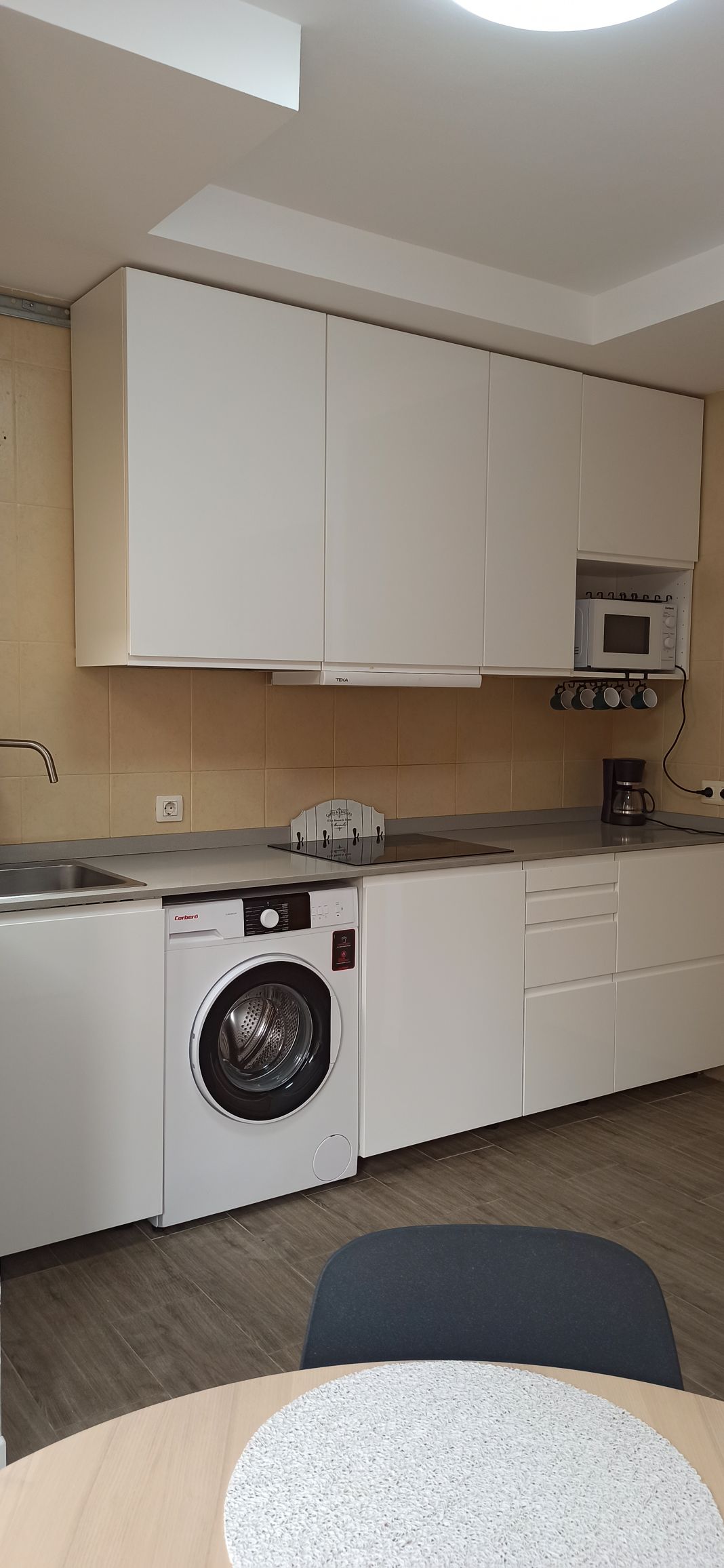 A functional kitchen is equipped with modern appliances, including a washing machine and a microwave. White cabinetry offers ample storage. A stainless-steel sink is positioned beneath a wall-mounted shelf, which holds coffee tools. Smooth countertops provide a clean workspace, complemented by a dining table and chairs.