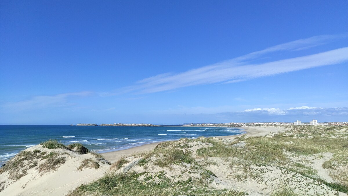 A sandy beach is visible along a tranquil coastline, with gentle waves lapping at the shore. The sky is clear, showcasing a vibrant blue hue, while wispy clouds drift above. The distant outline of buildings and land can be seen along the horizon.